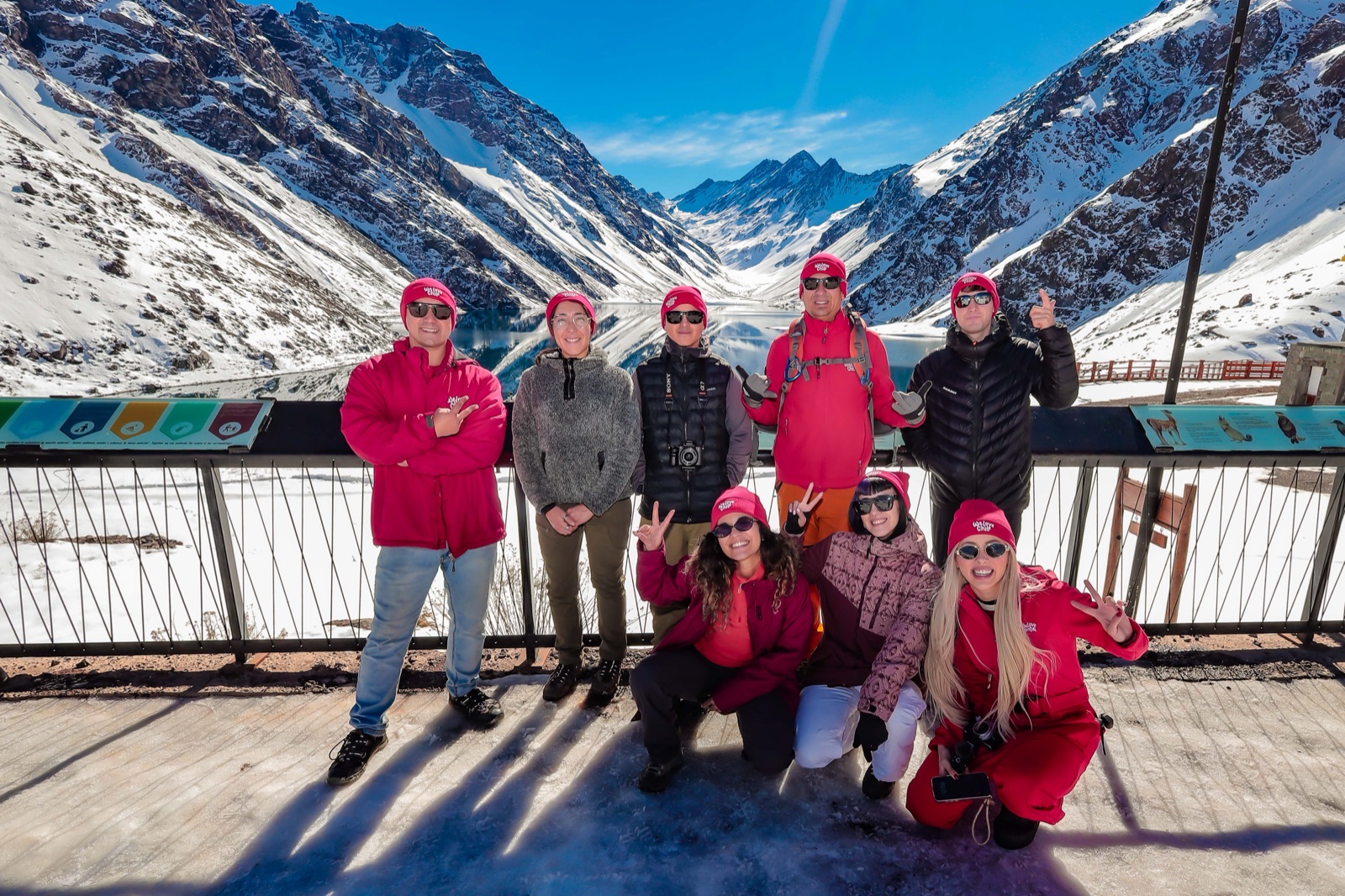 Grupo da We Love Chile de gorro e jaqueta vermelhos, posando diante da Laguna del Inca cercada de montanhas nevadas.