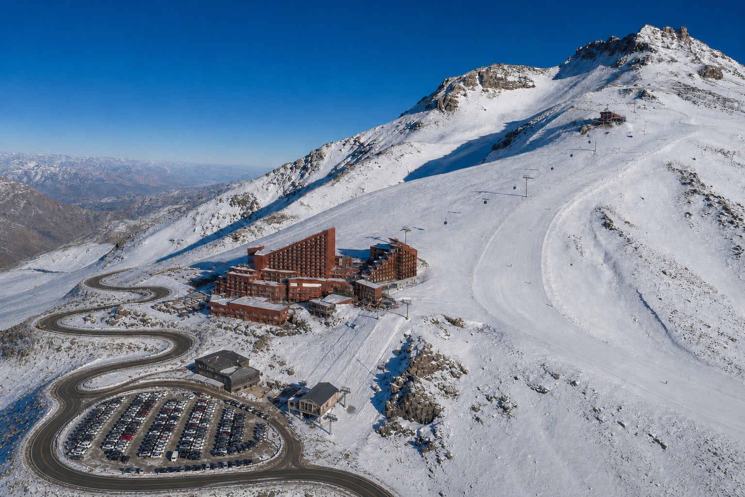Foto aérea da encosta do Valle Nevado no inverno: o estacionamento e o lodge da base a 3.000 metros, o complexo hoteleiro a 3.025 metros, a gôndola vermelha subindo até o Bar Bajo Zero a 3.200 metros e a pista mais alta a 3.670 metros.
